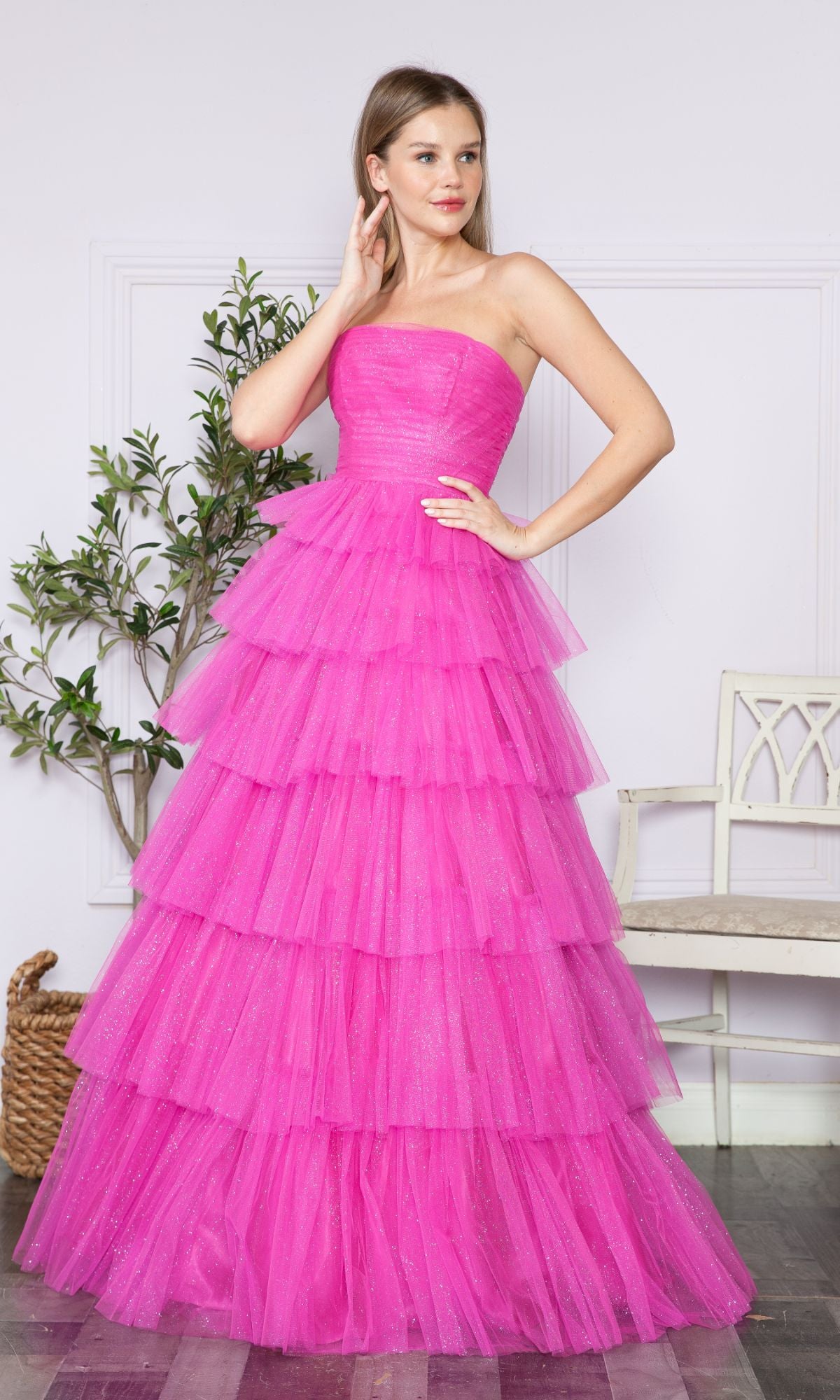 A woman stands indoors wearing the Strapless Long Tiered Glitter Prom Ball Gown 9386 in bright pink tulle, with a plant and a white bench in the background.